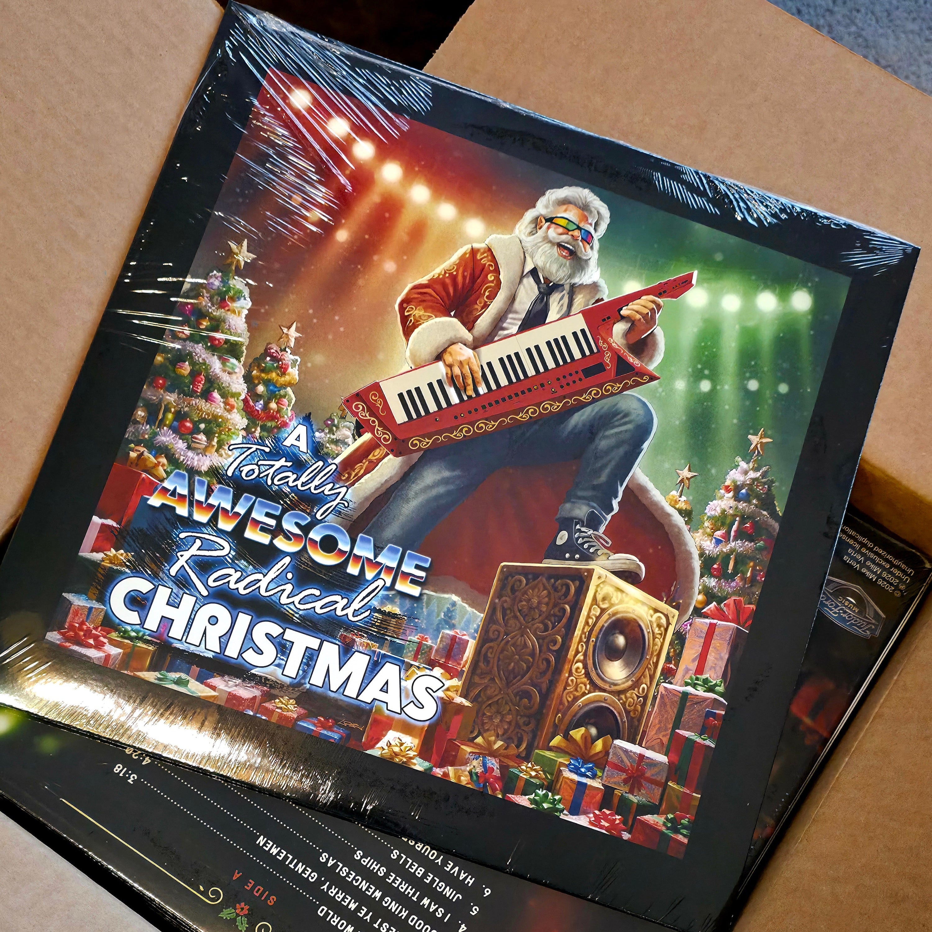 A colorful vinyl record wrapped in clear plastic, shown inside an open cardboard shipping box. The cover art features a rockin' Santa Claus playing a red keytar surrounded Christmas trees, gifts, and stage lights, with the text 'A Totally Awesome Radical Christmas' .